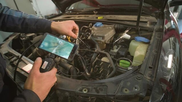 Hands of car mechanic check the vehicle engine with technical endoscope with rotary camera and mobile phone. Repair of engine head and valves.