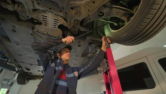 Young professional mechanic man working under lifted modern car in auto repair service. Car service, repair maintenance.