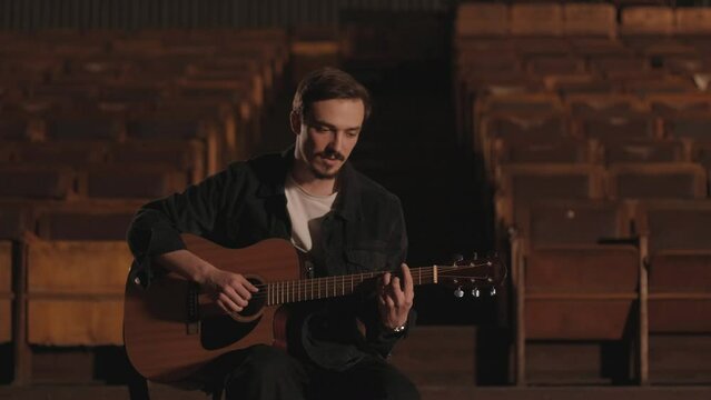 A handsome guy plays an acoustic guitar in an abandoned cinema. The musician sings a song and accompanies on the guitar