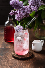 Refreshing cold lilac lemonade in the making. Sparkling soda water, lilac syrup, flowers, highball glass filled with ice cubes.
