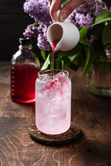 Refreshing cold lilac lemonade in the making. Sparkling soda water, flowers. Adding lilac syrup into a highball glass filled with ice cubes.