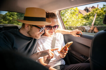 Young cheerful happy couple of travelers sitting in car looking at smartphone screen, travel comfort safety taxi