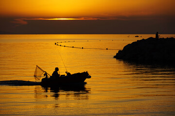 Golden hour sunset on seascape with black silhouette of fisherman in boat, stony pier and secure net on the sea surface