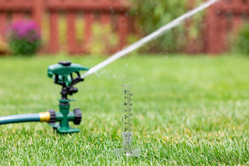 Rain gauge with lawn sprinkler watering grass in yard. Drought, water conservation, restrictions and lawncare concept - obrazy, fototapety, plakaty