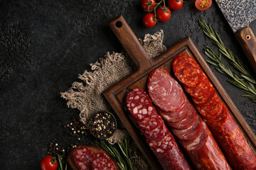 Sausage and cold cuts. Types of different loaf sausages on an old wooden board on black background and burlap with cherry tomatoes, rosemary, garlic and spices.Rustic.Top. Background image, copy space