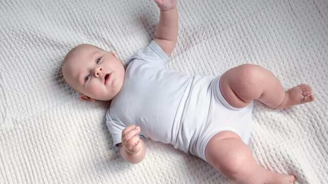 Beautiful Newborn Baby lying on his back on white blanket. Small Child wearing white pants and blue bodysuit. Cute little Baby is looking to Camera and gesturing. Childhood, Health Concept. 4K