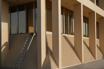 Corner of a nondescript yellow office building with pillars, windows, and a ladder. Has pleasing lines, textures, and shapes. Generic and minimalist. Good for backgrounds and horizontal copy space.