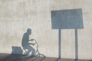 Shadow of delivery man on bike passing through a shadow of a rectangular street sign cast on a concrete wall. As a concept: the everyday cycle of making a living. For ads and horizontal copy space.
