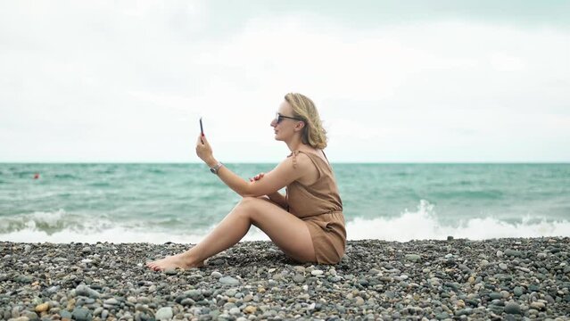 Girl talking on video call sitting on the beach on the background of the ocean with waves.