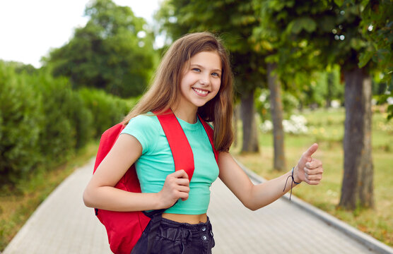 Outdoor portrait of cheerful school child. Happy beautiful teen Caucasian student girl in casual green T shirt with red backpack standing on park path, looking at camera, giving thumbs up and smiling