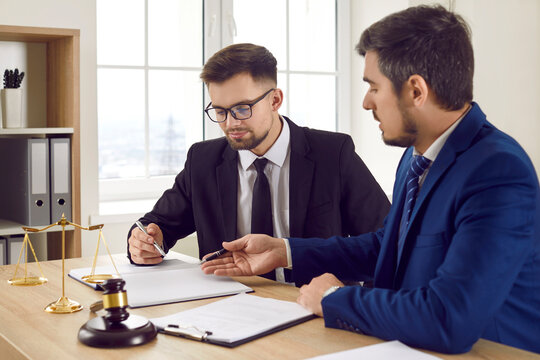 Two professional male lawyers read legal documents and study information about court case together. Men in suits sit in office at table near judge gavel and scales of justice and discuss legal issues