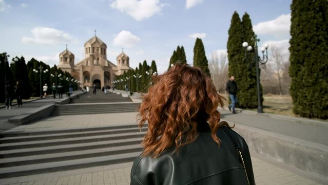 Travelling woman walking towards Saint Gregory the Illuminator Cathedral. Action. Yerevan Cathedral and stairs decorated by growing tujha trees on blue cloudy sky background.