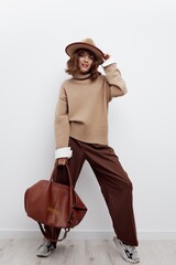 a beautiful woman stands full-length on a light background in a hat, sweater and shirt, sat down a little with her legs wide apart, holding a bag in her hand and touching the hat