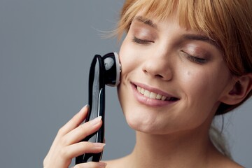 a woman with perfect skin on a gray background wrapped in a towel and does a facial massage with a black electric anti-aging massager, smiling pleasantly with her eyes closed. Close horizontal photo