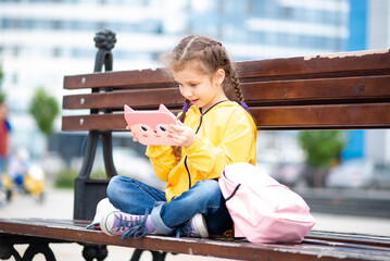 A girl of 6-7 years old holding tablet computer in her hands. She sits outside on a bench....