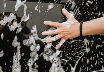 A male professional worker, a washer put his hand on the black dirty surface of a car door and white soap suds. Photography, concept.