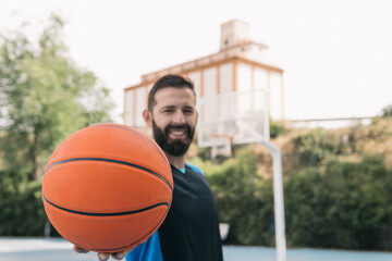 Smiling bearded young guy offering the ball is a blue street basketball court. Frontal image of a sporty man with a sleeveless shirt showing an orange ball with one of his hands. Sport concept