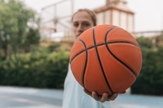 American blonde girl offering a basketball on the court. Close-up image of a sporty blurry woman displaying an orange ball challenging her opponent to start a street basketball game. Sport concept