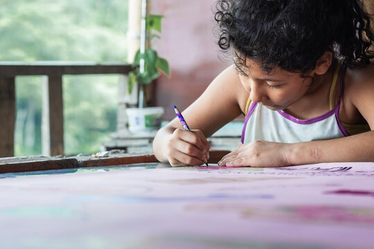 poor latina girl with brown skin very concentrated painting her drawing. disheveled girl with frizzy hair and old clothes doing her homework at a desk next to nature. concept of poverty and humility.