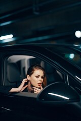 Fototapeta na wymiar a vertical photo from the side, at night, of a woman sitting in a black car and looking out of the window and gently touching her face while adjusting her makeup looks into the side view mirror