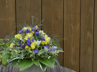 Strauss aus echten Blüten mit blauen Glockenblumen und gelben Rosen, weissen Matrikaria, Leinkraut und Funkienblättern vor Hintergrund aus Holzdielen