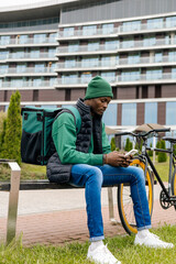 A young black delivery man in a city park on a bench takes an order on his phone.
