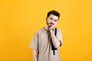 Caucasian man wearing beige tee posing isolated over yellow background feeling bored and tired. depressed guy touching his cheek with a palm. Leaning head on palm. Depression concepts.