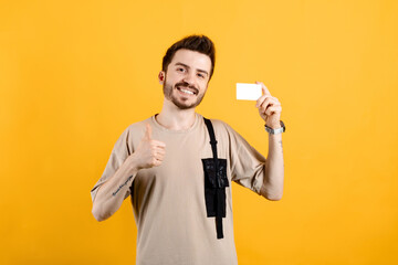 Cheerful young man wearing t-shirt posing isolated over yellow background Holding credit bank card showing thumb up. Mock up copy space.