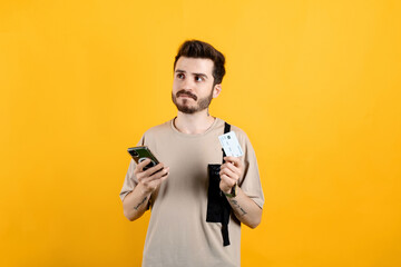 Handsome caucasian man wearing beige t-shirt posing isolated over yellow background holding smartphone and credit card with anxious expression while looking aside.
