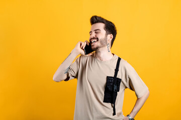 Happy young man wearing beige t-shirt posing isolated over yellow background talking on smart phone and smiling. Receiving good news during phone conversation.
