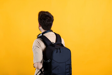 Handsome caucasian man wearing beige t-shirt posing isolated over yellow background student wearing a backpack with headphones around his neck. Back view photo. - obrazy, fototapety, plakaty