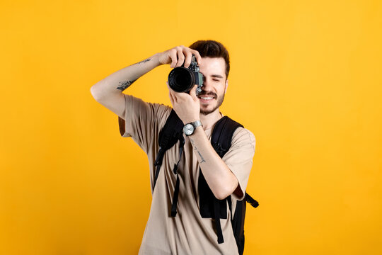 Young man smiling confident wearing beige tee posing isolated over yellow background taking images with dslr camera. Photographer covering his face with the camera.