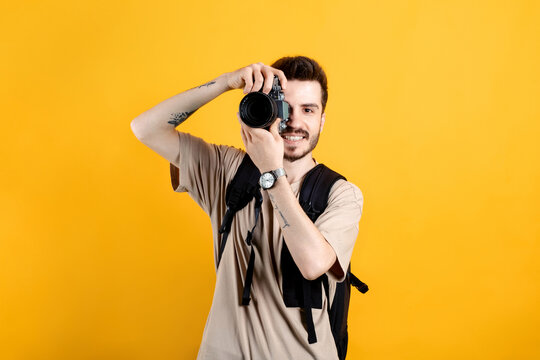 Caucasian man wearing beige tee posing isolated over yellow background taking images with dslr camera. Photographer covering his face with the camera.