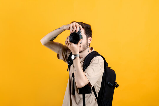 Caucasian young man wearing t-shirt posing isolated over yellow background taking images with dslr camera. Photographer covering his face with the camera.