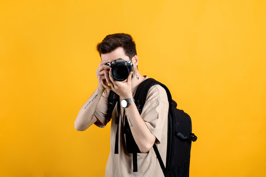 Handsome caucasian man wearing beige t-shirt posing isolated over yellow background taking images with dslr camera. Photographer covering his face with the camera.