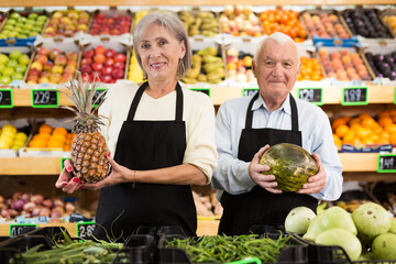 Two supermarket workers, old man and woman standing in salesroom with pineapple and melon in hands.