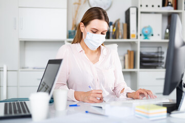 Businesswoman in protective mask working alone with laptop and papers in office, new normal due to...
