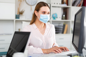 Female office worker in protective medical mask is having productive day at work in office