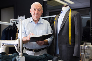 Elderly man tailor takes measurements from his jacket and writes them down in notebook in a workshop