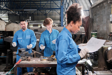 Young African American female industry worker works with mechanical drawing and metalwork precision tools, lathe machines, and spare parts workshop with multiracial team in a manufacturing factory.