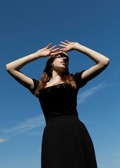Fototapeta na wymiar an elegant woman in a black dress stands against the blue sky in sunny weather covering her face with her hands