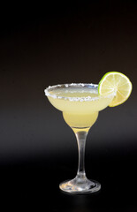 Margarita cocktail with a slice of lime and salt around the edge of the glass on a black background.