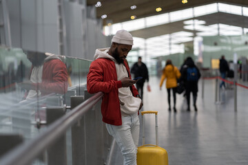 Thoughtful African American traveler man using smartphone at airport terminal, travelling with luggage bag. Passenger guy waiting her flight, checks time of flight online, standing in railway station