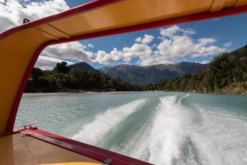 The wake behind a speeding jet boat on the waters of the lower Waiatoto River surrounded by thick rainforest, on a sunny day. West Coast, New Zealand.