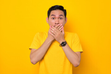 Shocked young Asian man in casual t-shirt covering mouth with hands and looking at camera isolated on yellow background. People lifestyle concept