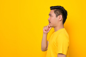 Thoughtful young Asian man in casual t-shirt looking aside at copy space isolated on yellow background. People lifestyle concept