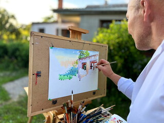 adult male artist 45-55 years old paints a picture looking at his house in front of him a sheet of paper on which you can already see the windows and green wood as well as many paints and brushes.