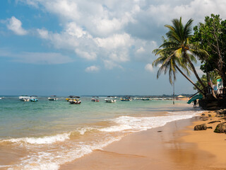 Hikkaduwa, Sri Lanka - March 8, 2022: Beautiful view of Hikkaduwa beach with green palm trees. Boats stand along the coast of the Indian Ocean