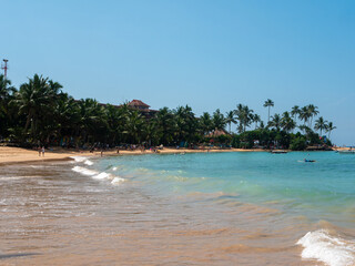 Hikkaduwa, Sri Lanka - March 8, 2022: Beautiful view of Hikkaduwa beach with green palm trees against the blue sky. People relax, sunbathe and swim in the azure water of the Indian Ocean. Copy space