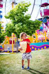 Cute bi-racial little boy eating a big bag of popcorn at an outdoor carnival. Smiling and having fun by a large ferris wheel at an amusement park